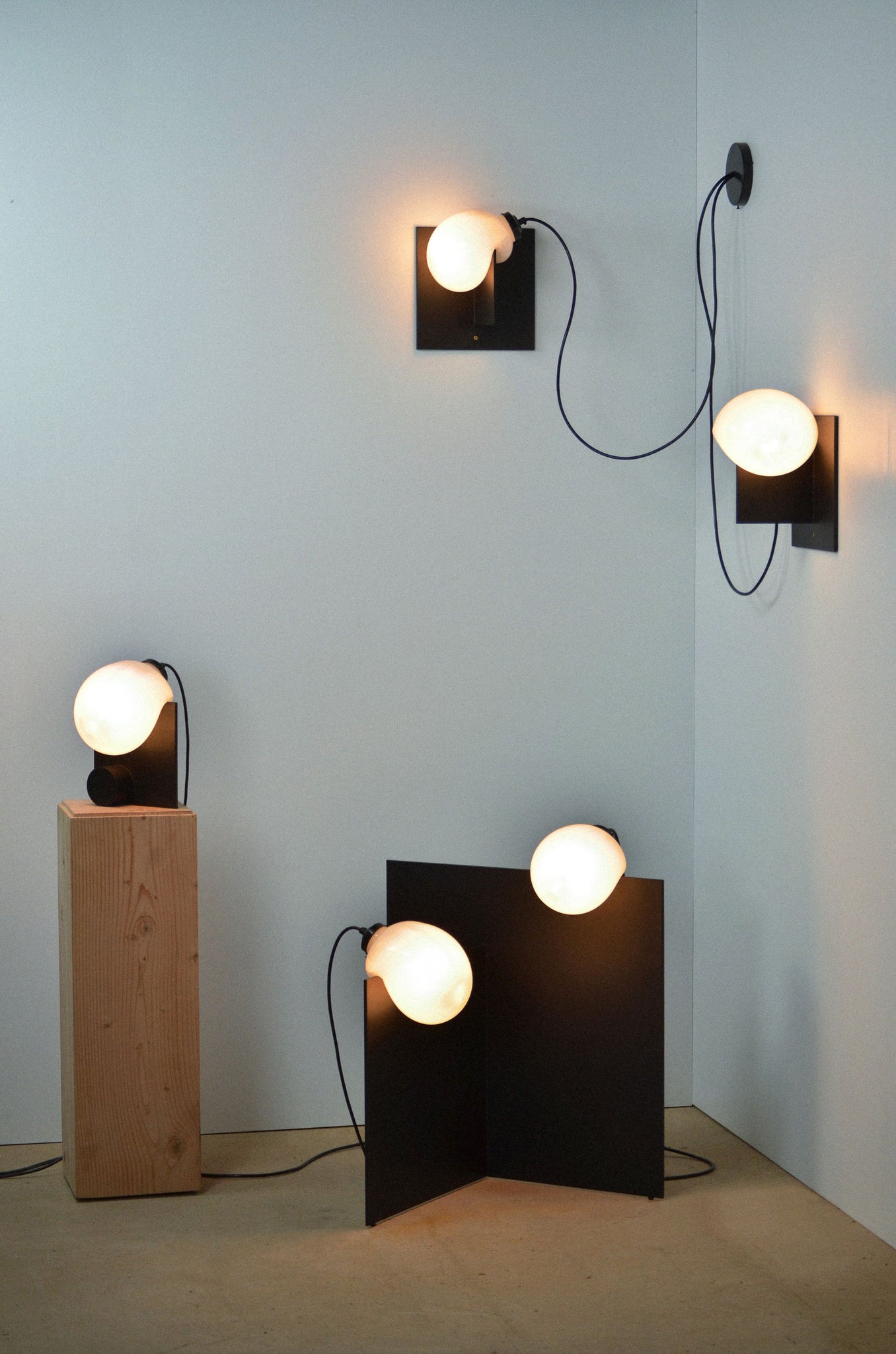 The Bloop Floor Lamp, Bloop Table Lamp on a wood pedestal, and two Bloop Sconces mounted on the wall in the corner
